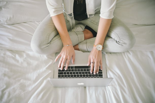 A woman wearing a large watch types on a Mac laptop while sitting cross-legged on a white bed. 