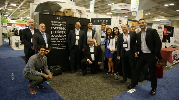 Eleven attendees gather smiling in front of the Luxer One package lockers booth.